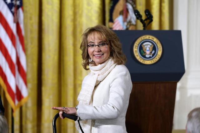 A white woman with shoulder-length dirty blonde hair and black glasses stands in front of a podium with a United States seal on it and an American flag to the left. She is wearing a white jacket and leaning on a cane that shows up in the bottom of the frame.