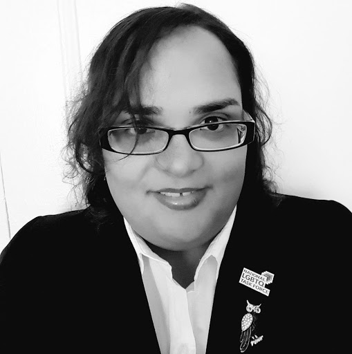 Black and white photo of a Latina woman with dark hair and black rimmed glasses smiling for the camera. There's a small gap in her front teeth and she is wearing a blazer with multiple pins down one lapel, including one that says "National LGBTQ Task Force."