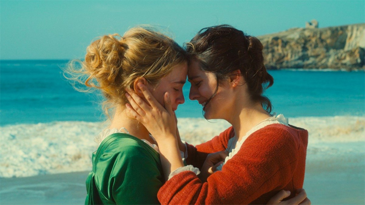 A screenshot of "Portrait of a Lady on Fire" in which one woman holds another woman's face on a beach. This is a lesbian movie available for streaming on Hulu.