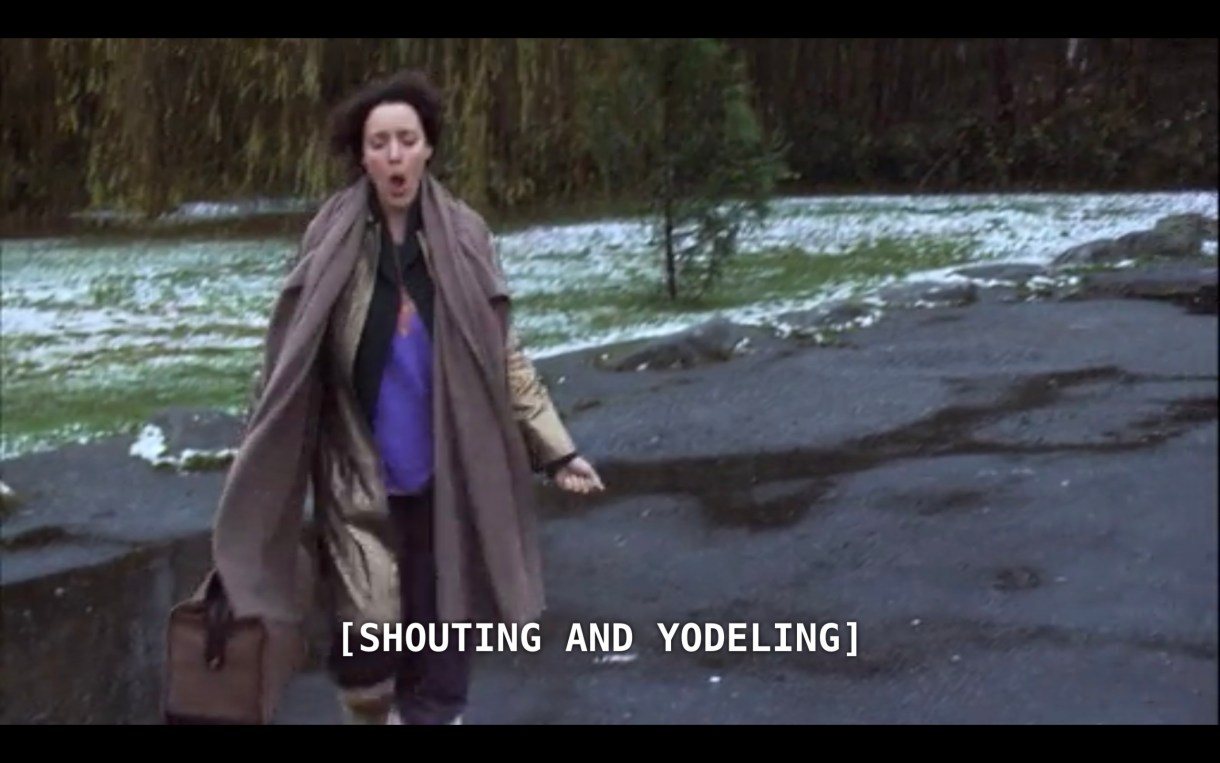 Bette (wearing a purple shirt under a long brown jacket) is carrying a duffle bag as she walks happily down a lightly snowy road as she continues to leave her silent retreat. The subtitles show that she is "[shouting and yodeling]"