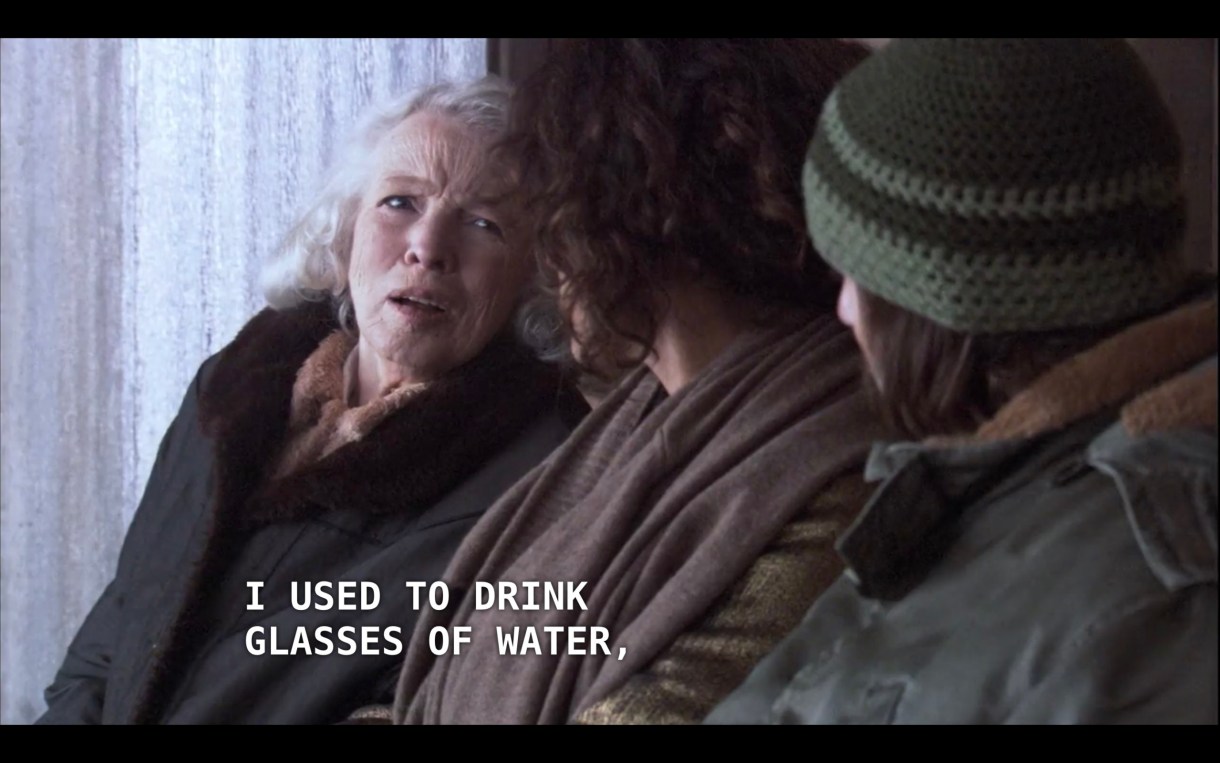 Bette sits in between two strangers at a bus stop. On her right is an older woman with white hair and wearing a black coat. On her left is a guy with a green knit hat and green jacket. The woman says to Bette, "I used to drink glasses of water"