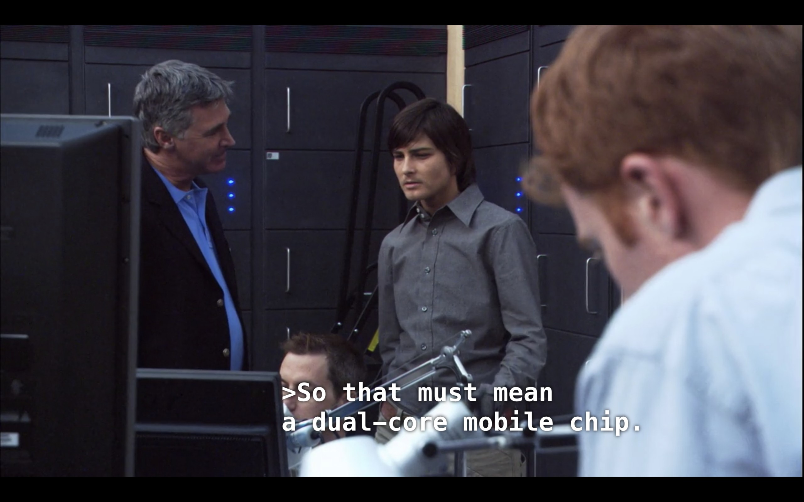 Max (wearing a grey button-up) is standing in a tech office with three other white guys in suits. Max is looking at a computer monitor and says, "So that must mean a dual-core mobile chip"