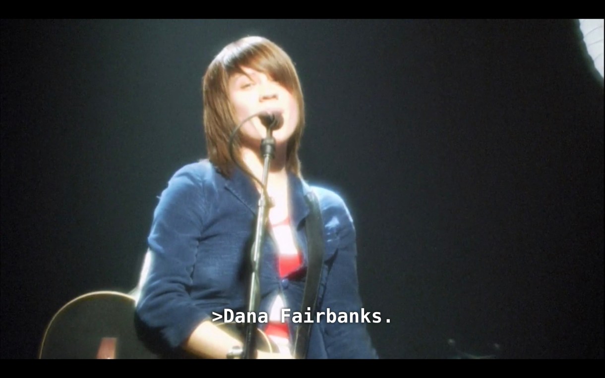 Tegan (or Sara? Hard to say) Quin is wearing a blue blazer and standing at a microphone on stage. She responds to her sister's question, saying, "Dana Fairbanks."