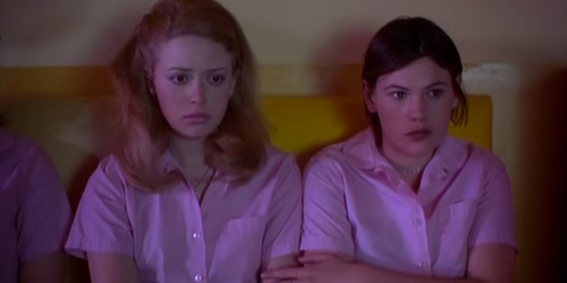 A still from the best lesbian movie of all time But I'm a Cheerleader. Two girls watching a movie in pink outfits.