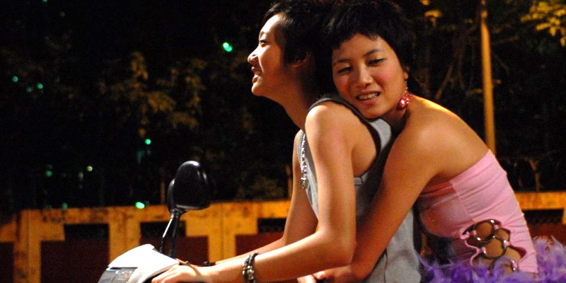 A femme rides on the back of a motorcycle with her arms wrapped around a masc. 