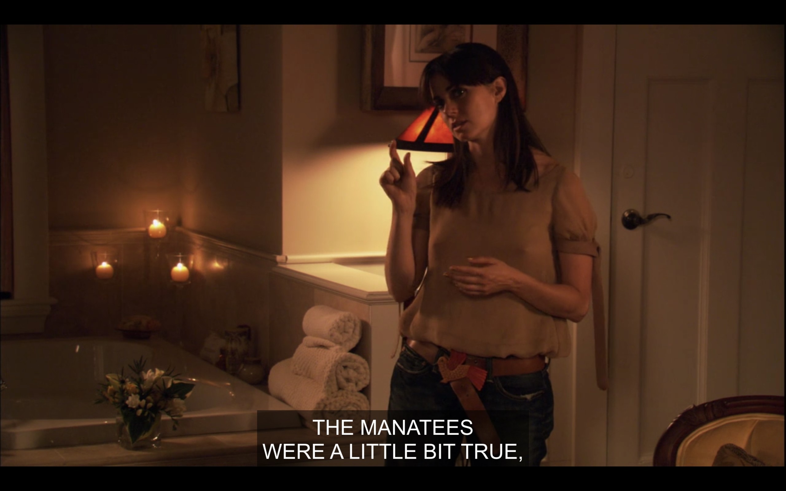 Jenny (wearing a peach colored top) stands in a dimly lit bathroom, with lit candles and flowers by the bathtub. She says, "The manatees were a little bit true."