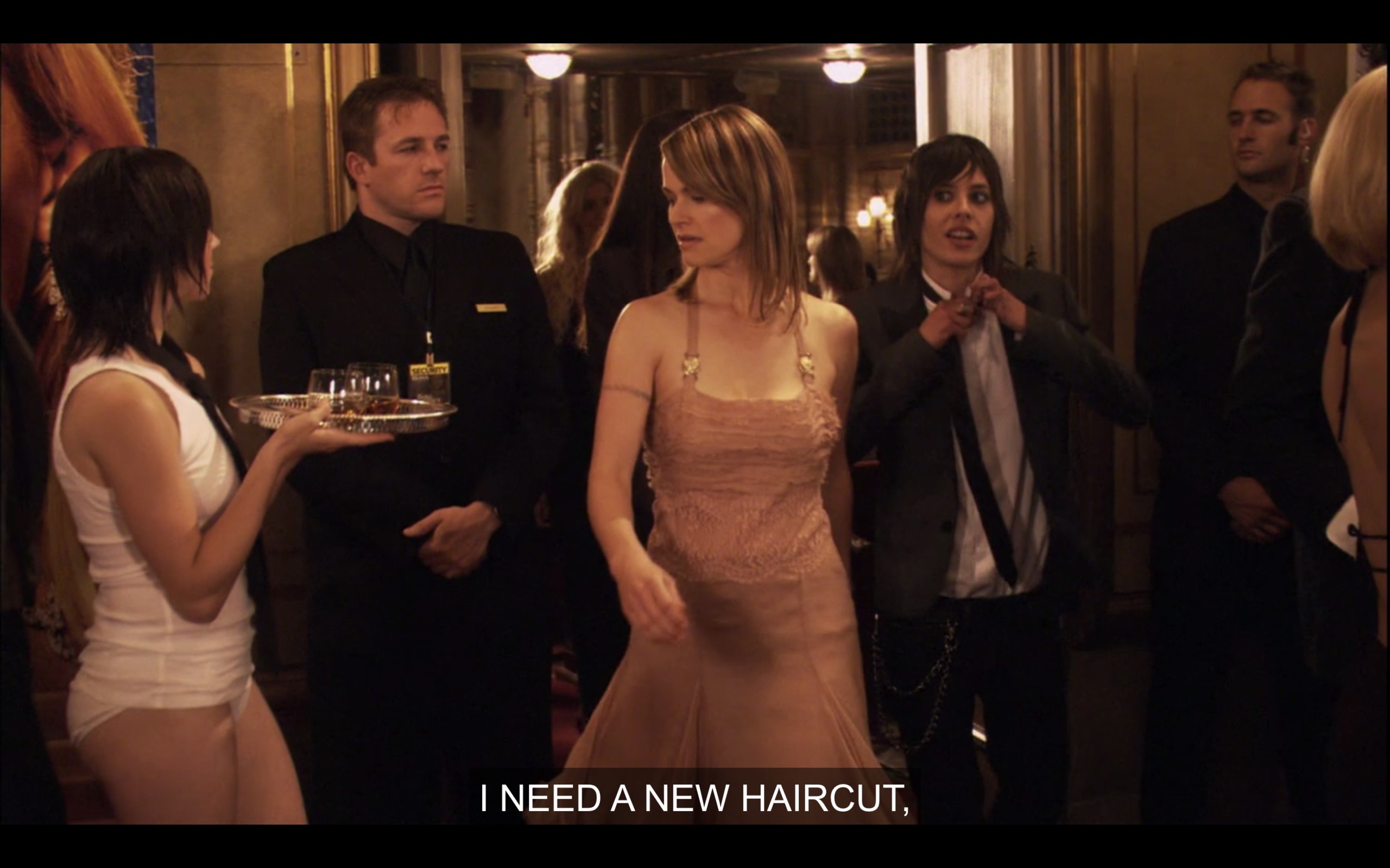 Alice (wearing a peach dress) and Shane (wearing a skinny black tie, white shirt, and black blazer) pass a caterer (wearing white brief underwear and a white tank top only) carrying a plate of food. Shane is loosening her tie and says, "I need a new haircut."