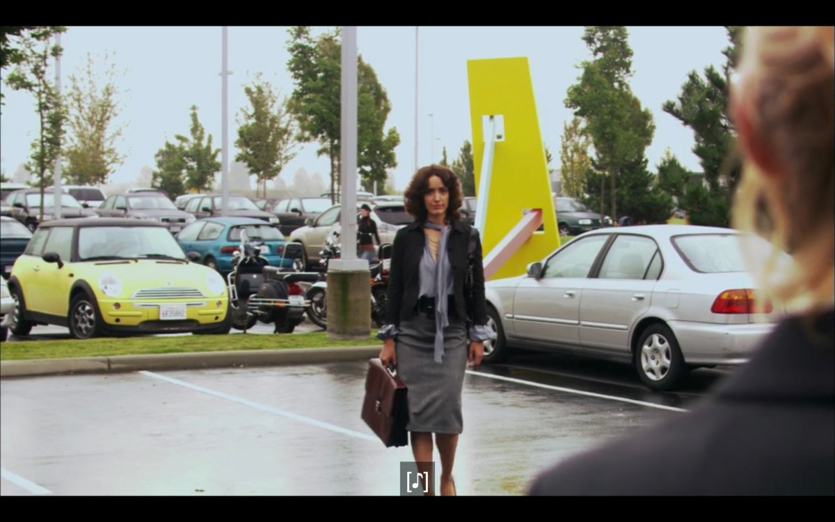 Bette (in a grey skirt, black jacket, carrying a brown brief case) walking through a parking lot towards Tina (who is barely in frame).