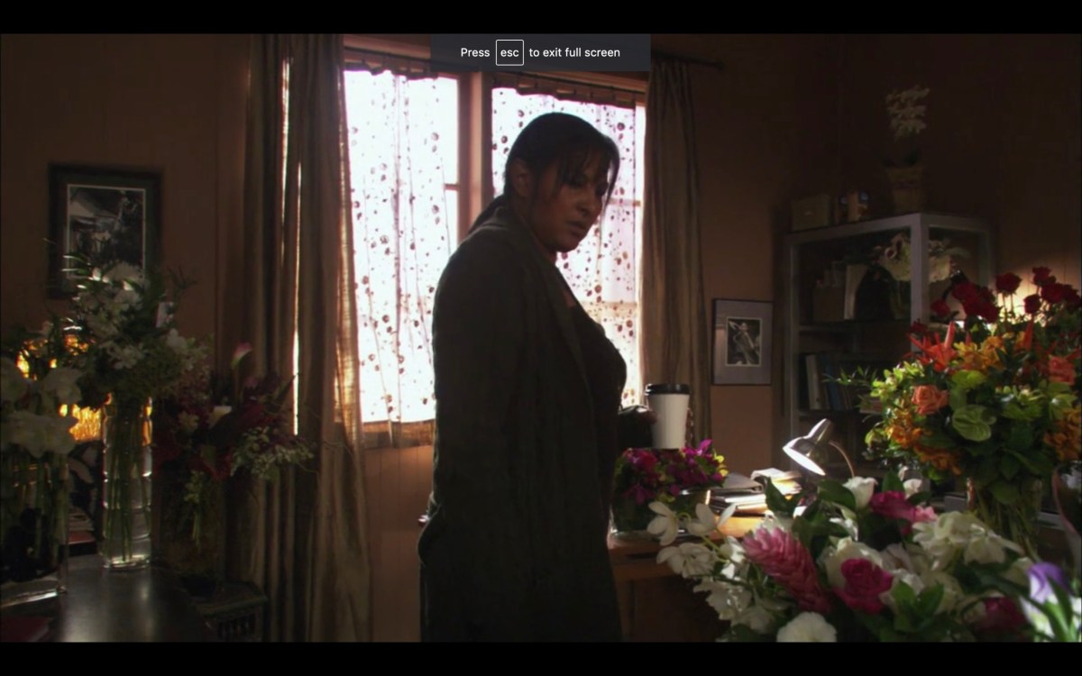 Kit in a room full of flower bouquets. She is holding a white disposable coffee cup. She is looking quizzically at the flowers.
