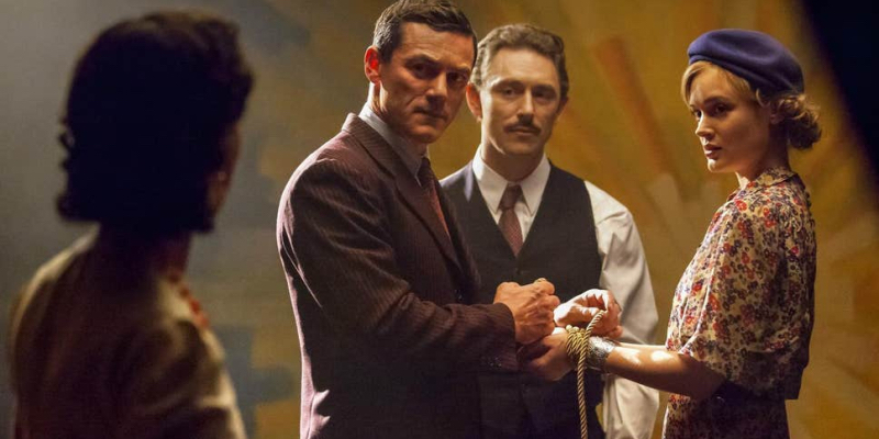 A still from the 16th best lesbian movie of all time Professor Marston & the Wonder Women. A man demonstrates a rope tie on a woman's wrist while looking at another woman.