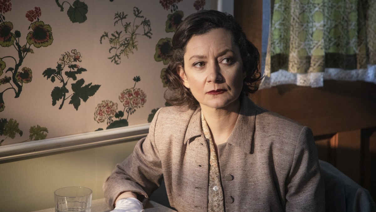 Sarah Gilbert as "JM," sitting in her home at a desk, looking uneasy