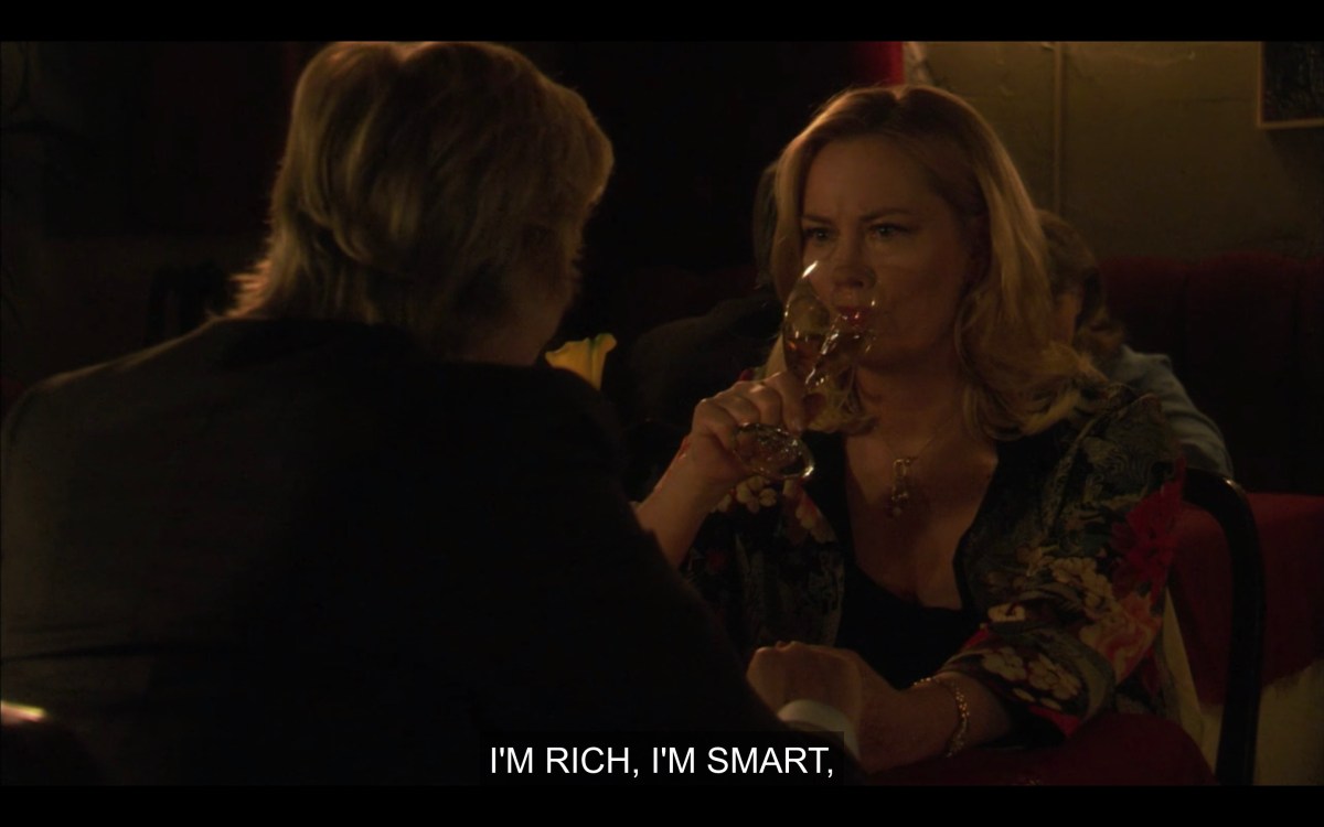 At a restaurant. Phyllis downing a glass of white wine while Joyce says, "I'm rich, I'm smart"