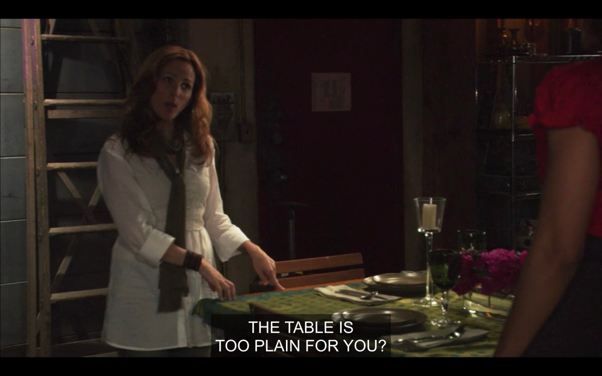 Jodi gestures towards the table in her loft, which is set with a green tablecloth and place settings. Jodi is wearing a white shirt and scarf and gesturing towards the table saying "the table is too plain for you?"