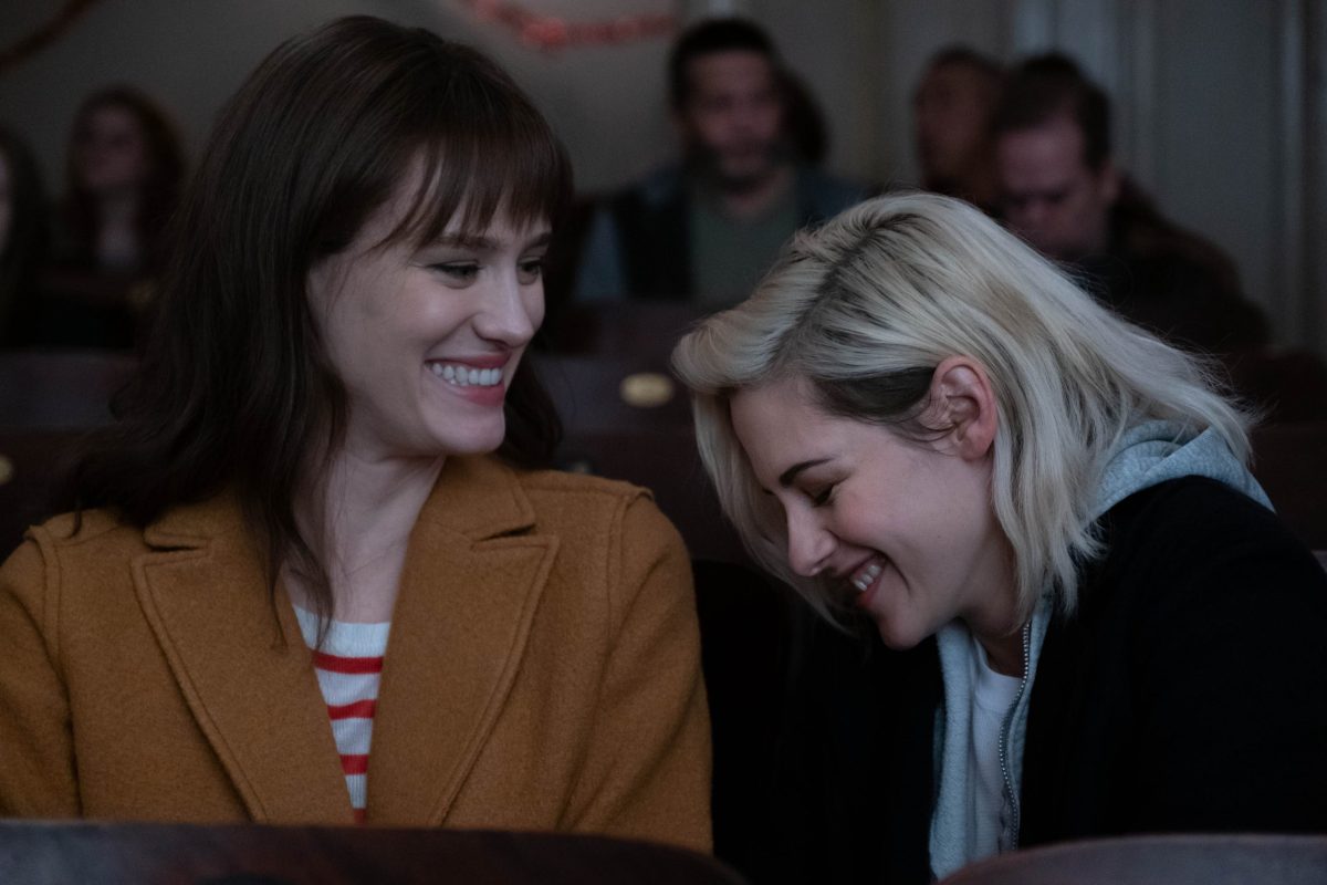 A screenshot of "Happiest Season" in which Kristen Stewart smiles at Mackenzie Davis. This is a lesbian movie available for streaming on Hulu.
