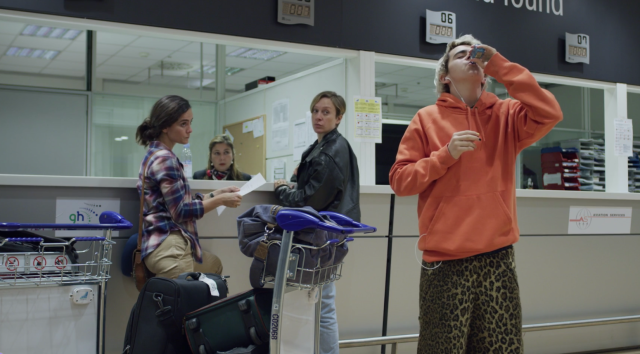 three women stand off to the side while a teenage boy in flashy clothing sucks down a bottle of alcohol.