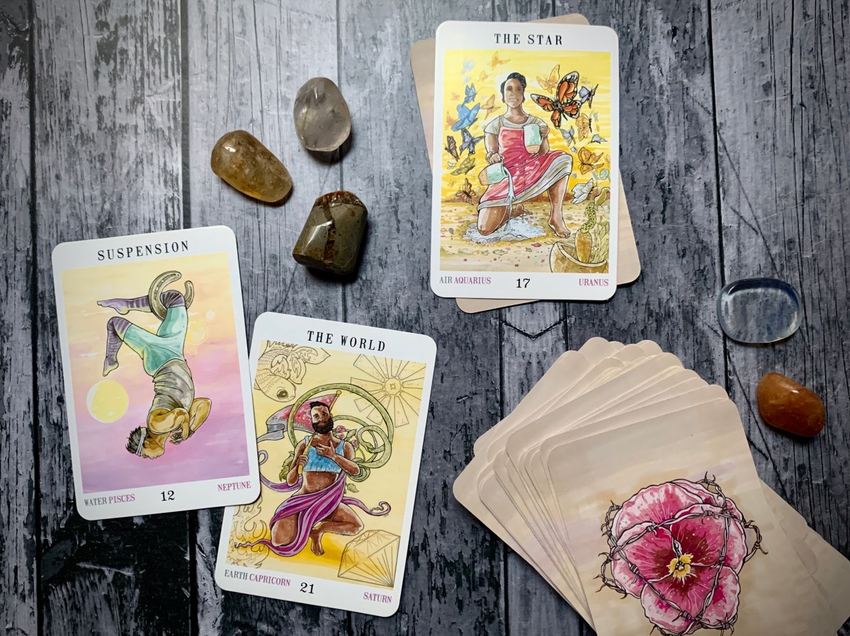 The Star card, the Suspension card, and the World card from the Next World tarot deck upturned on a wooden backdrop with a number of stones and crystals.
