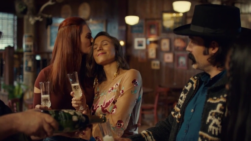 Nicole Haught gently kisses a smiling Waverly Earp's temple as they stand in Shorty's, Waverly with a champagne flute in her hand.