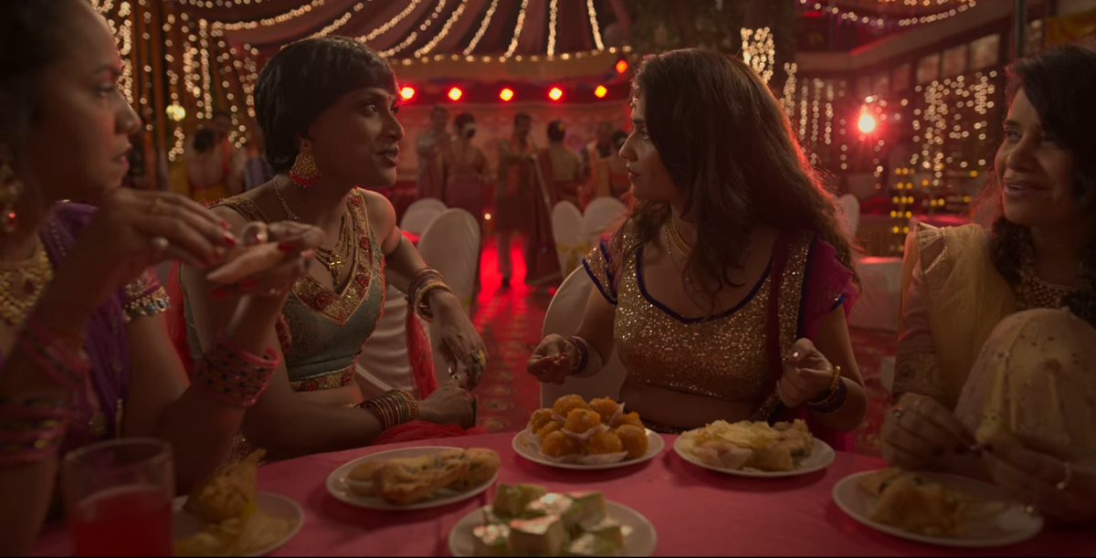 Bombay Begums queer storyline: Lily's friends are dressed in traditional Indian clothes and sit around a table full of sweets. Lily's friend Beauty is smiling and talking to Lily. Lily is surprised by Beauty's question. 