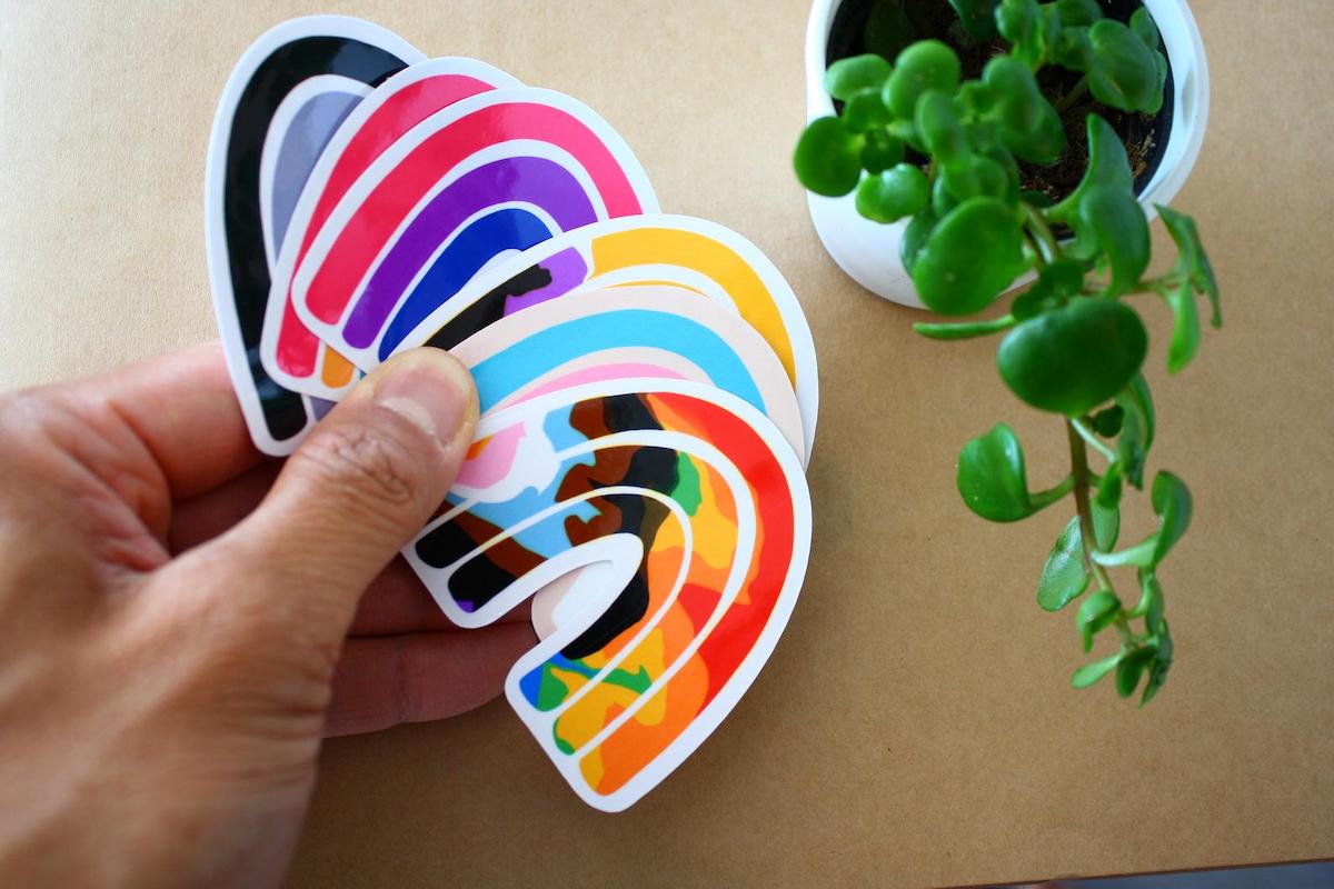 A person holds several rainbow stickers, some are for the trans flag, others are different colors.