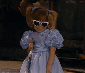 one of the olsen twins in a fluffy lavender dress holding flowers and wearing sunglasses moves her body back and forth, looks cute and mischieveous