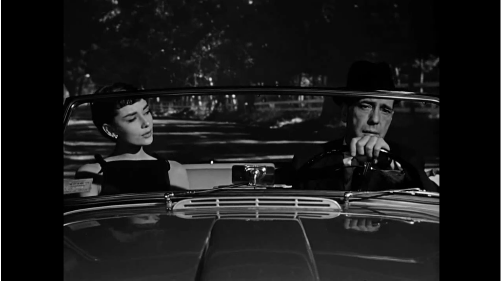 In the black-and-white movie Sabrina, Audrey Hepburn sits in a car next to Humphrey Bogart, who is driving