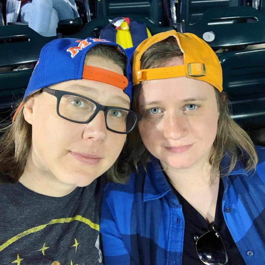 Heather and Stacy look like they are seated in some kind of sports stand. Heather and Stacy are white women with brown hair. Both are wearing backwards baseball caps. Heather is wearing glasses.