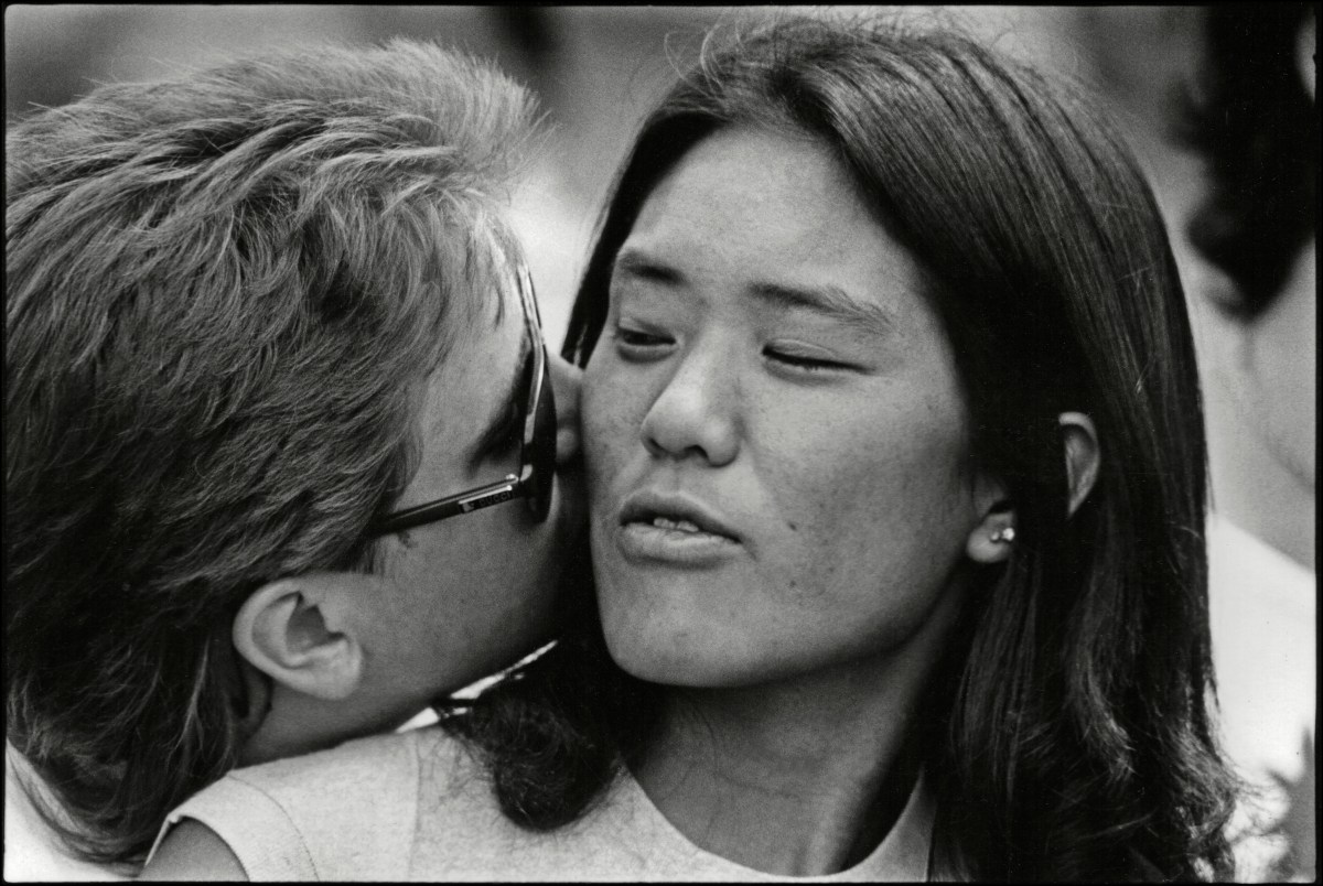 In a black and white photo, a white woman kisses the neck of an Asian woman.