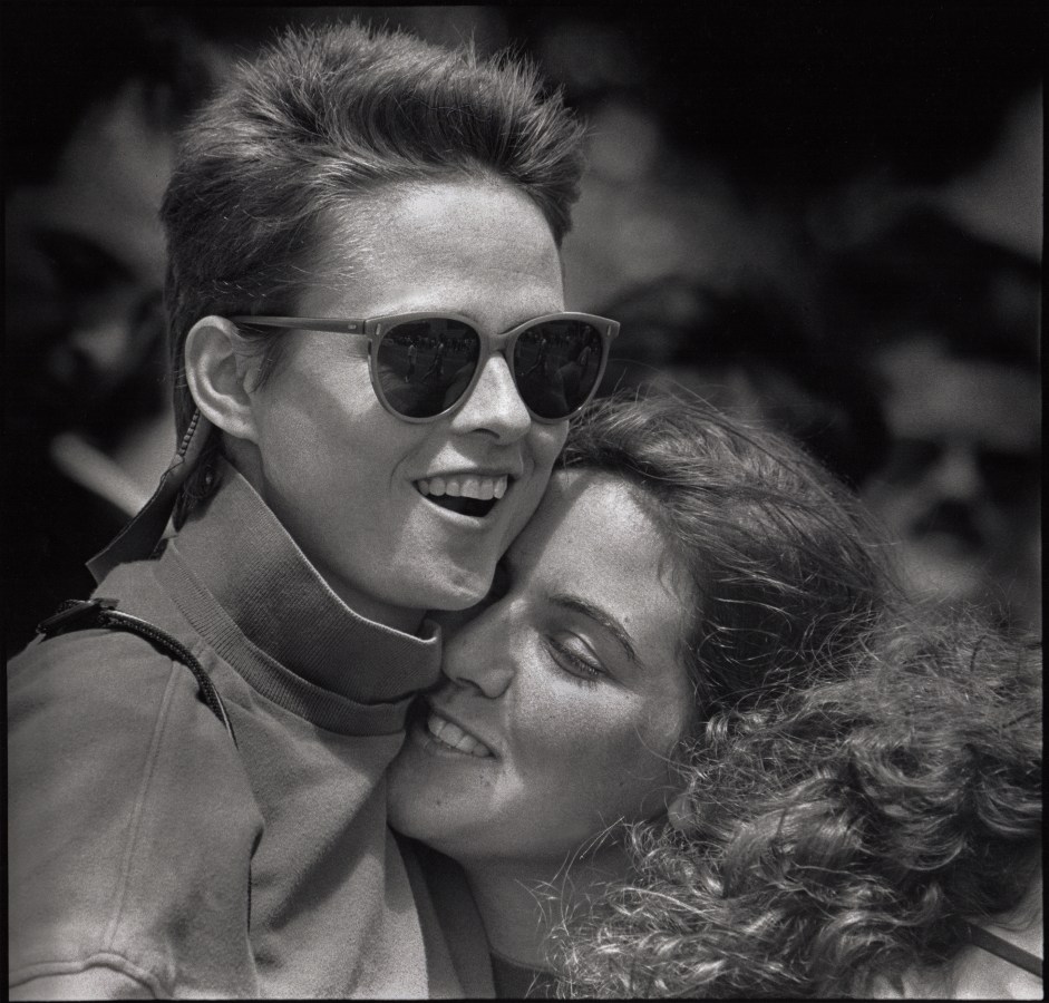 Close-up of two women as they embrace in a black and white photo, the taller woman has on sunglasses and the shorter woman has tucked her face into the neck of the taller woman.