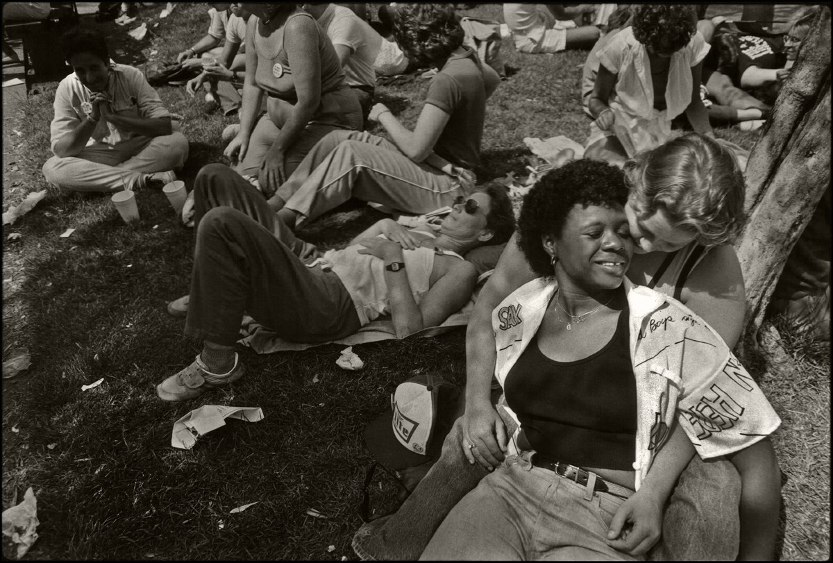 Among a crowd on the grass outside the Civic Center, two women share a smile together, one woman is black and lays across the lap of a woman who is white.