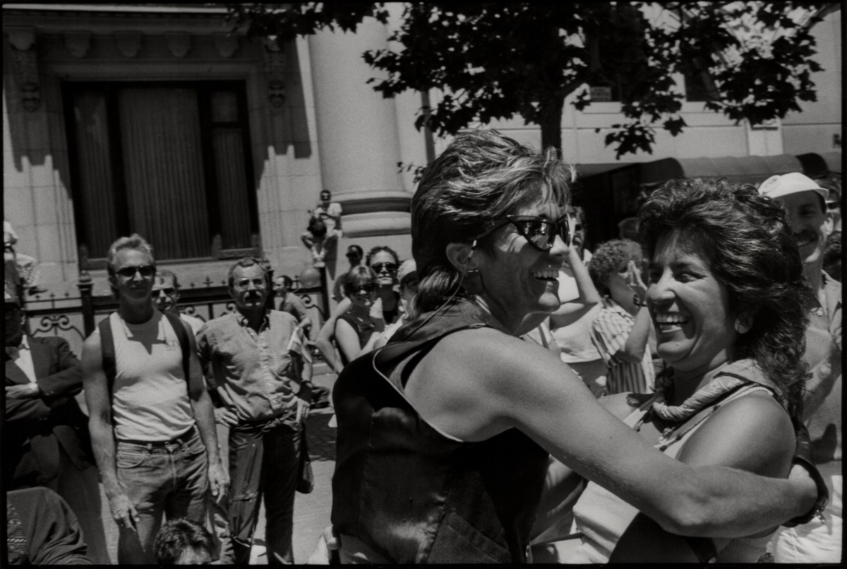 Two woman smile as they greet one another in a black and white photo, one woman has a leather vest and sunglasses and teased, short blonde hair. The other has fanned out brunette hair.