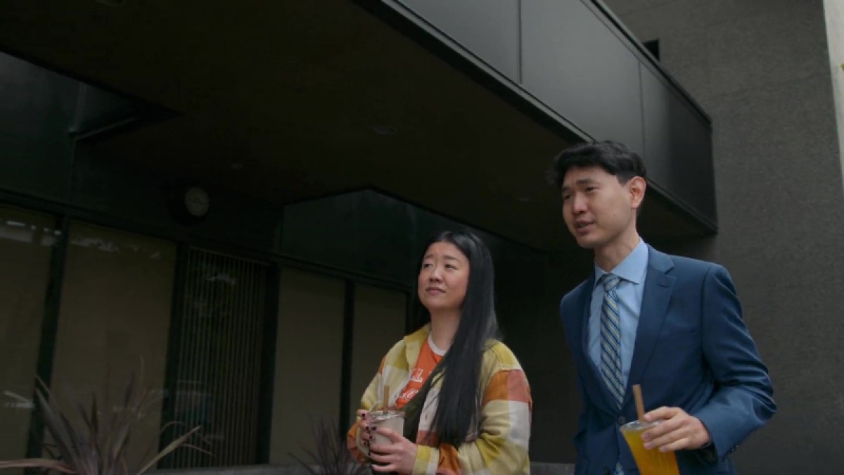 Alice and her brother spend time together this week on Good Trouble. He's wearing a dark blue suit with light blue shirt and tie, carrying an orange drink. Alice is on the left, wearing a flannel shirt over an orange t-shirt.