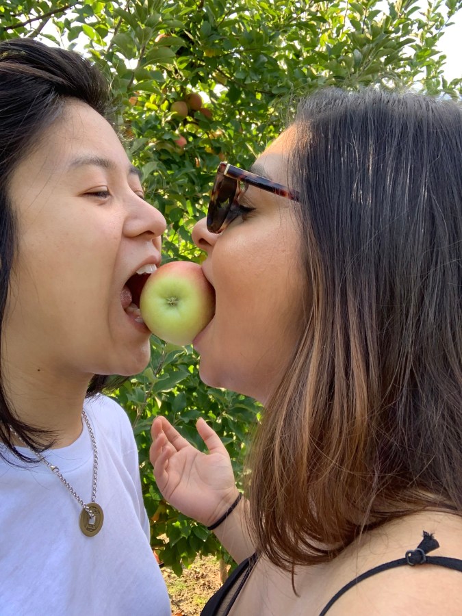 gerrie, a nonbinary Chinese-Filipino human, and Ashni, a South Asian gender averse human, each with medium length hair, gerrie's black and ashni's an ombre brown, bit down on an apple held between them by nothing but their mouths! ashni is wearing sunglasses. they look like they are having fun