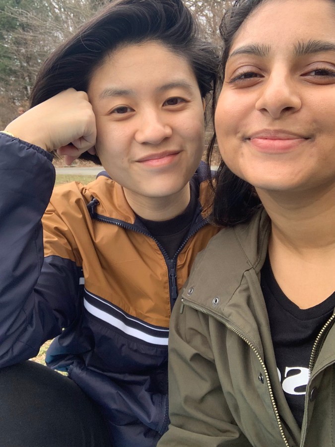 Gerrie and Ashni sit in what looks like a park, looking at the camera with satisfied smiles. They are both wearing light jackets.