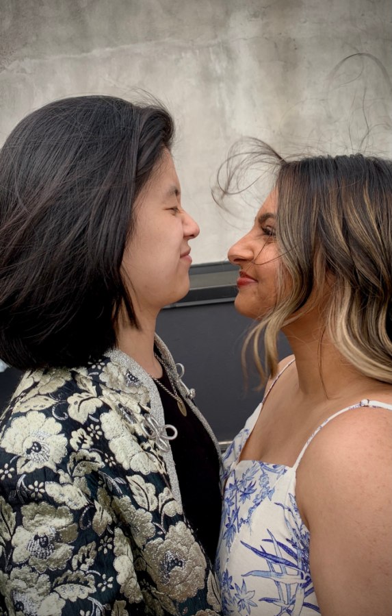 Gerrie and Ashni face off in this photo. Their hair is both a little wind blown. Gerrie has a straight black shoulder length cut and Ashni has a longer ombre situation. Gerrie is wearing a patterned jacket and Ashni a sundress.