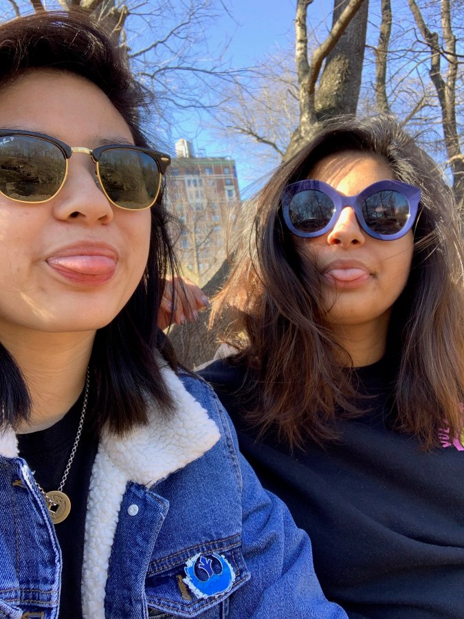 Gerrie and Ashni wear sunglasses outdoors. They both stick their tongues out playfully.
