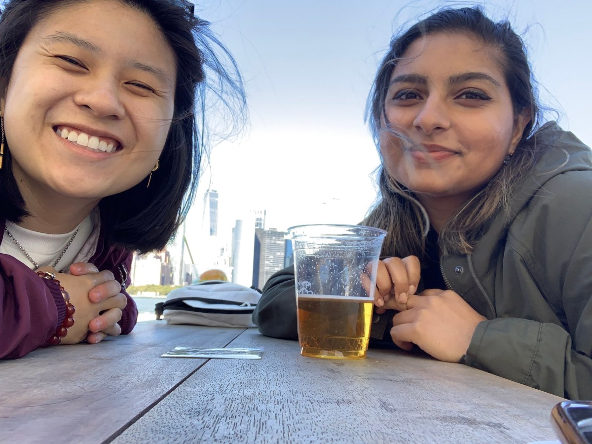 Gerrie and Ashni sit at a picnic table. It looks like Ashni has a beer in a plastic cup. Gerrie is smiling widely and Ashni is smiling with their mouth closed.