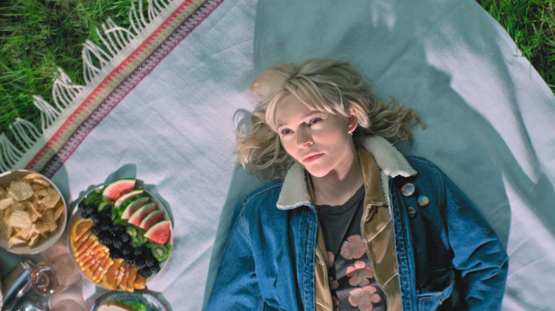 Raelle is lying on a picnic blanket in bright sunlight next to a plate of fresh fruit 