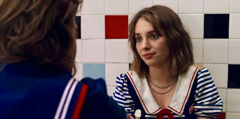 Robin from Stranger Things sits on the floor of the bathroom in her uniform from her job scooping ice cream, her mascara is smudged from crying