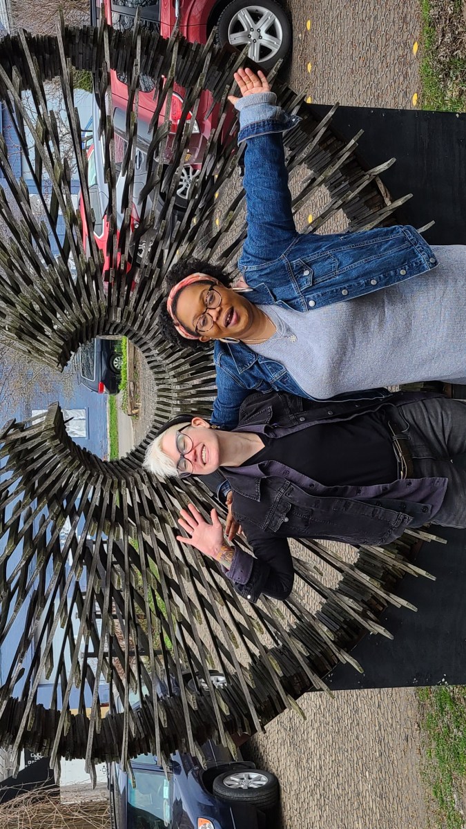 Carmen and Nico are hugging with their bodies open to the camera, they are in front of a metal sculpture that has an open circle with straight lines coming out. Nico is a white genderqueer person with blonde hair, a black denim coat, black shirt, and black jeans. They have on a black beanie. Carmen has on a pink headband, denim jacket, and grey sweater dress, and combat boots.