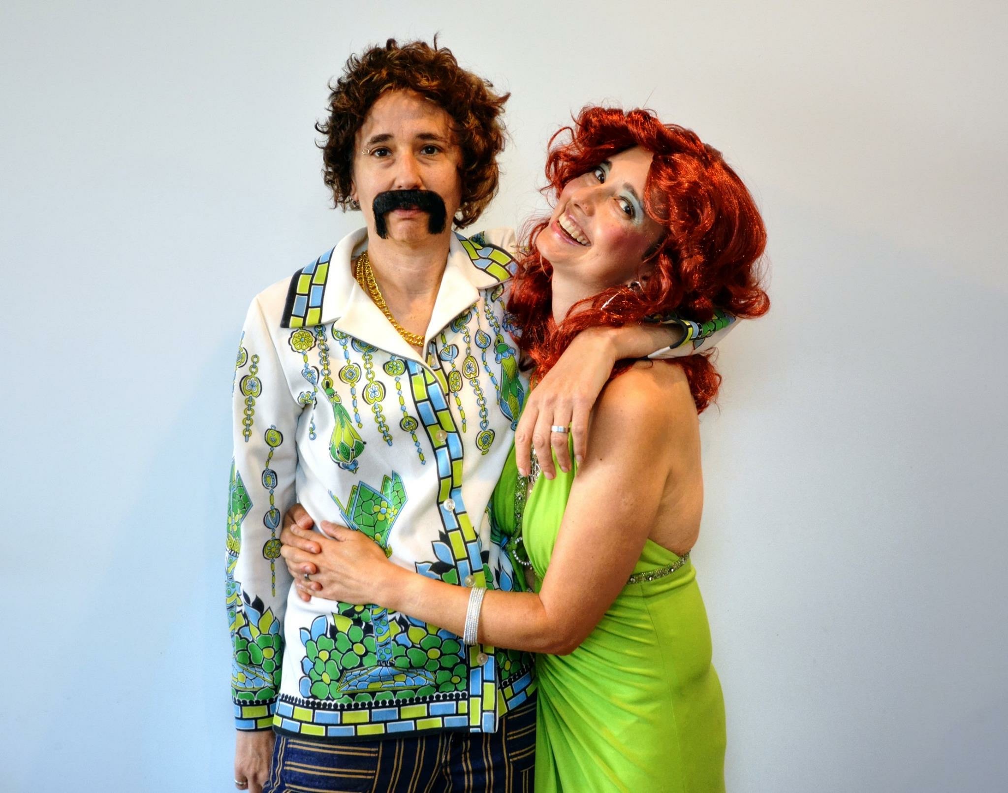 Tracy and Mia are dressed up for Halloween as a garish 1970s couple. Tracy wears a huge red curly wig and lime green dress and blue eyeshadow and his throwing her head back and laughing. Mia is wearing gold chains, a very loud button up shirt wiht green accents, and mis-matching striped pants. She also has a huge, fake handlebar mustache.