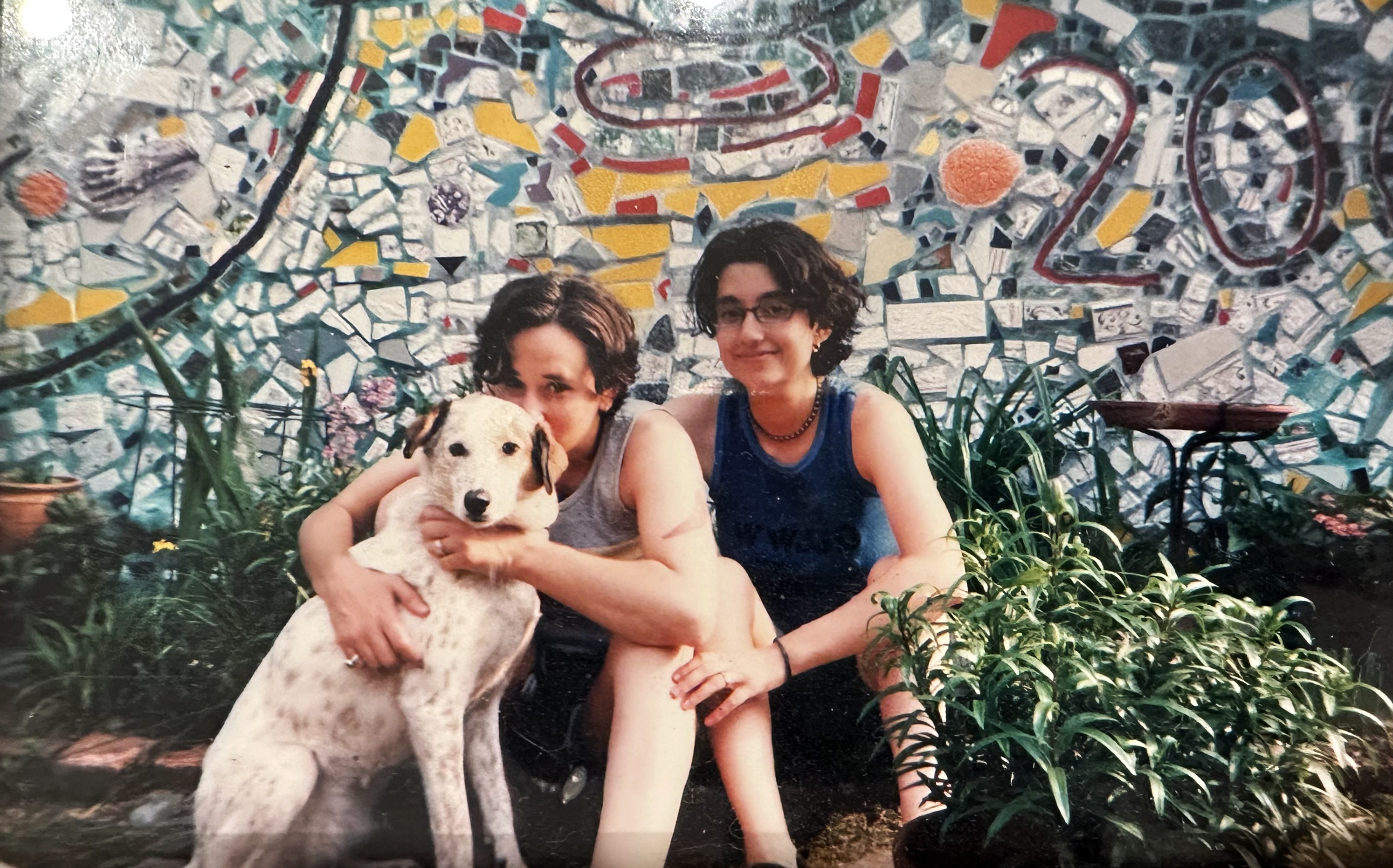 In this older photo, Tracy and Mia pose in front of a colorful mosaic while holding a very cute medium-sized dog.
