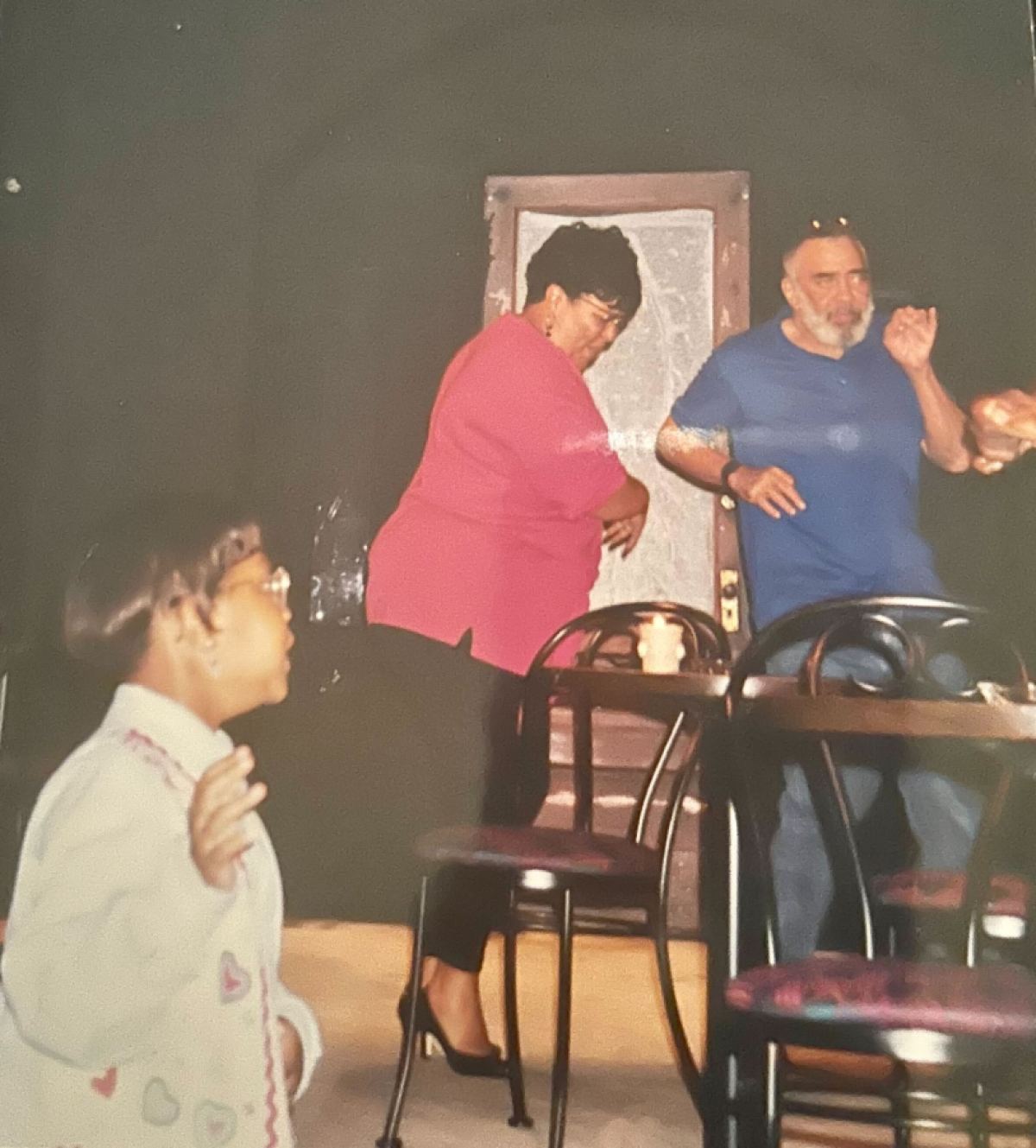 Carmen is in the blurry foreground of a photo of her mom at rehearsal, Carmen's mom is a light skinned Black woman in a pink shirt, black pants, and high heels. Carmen is in a denim button down shirt with pink hearts, her hair is cut into a mushroom cut and shinning in the camera.