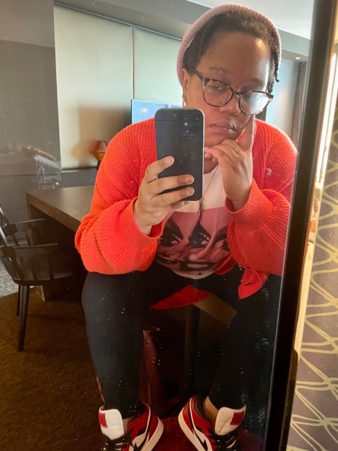 Carmen, a black femme, has a bright orange sweater and a purple beanie, black sweatpants, and red and black Jordans on her feet, she's leaning into a window and taking a selfie.