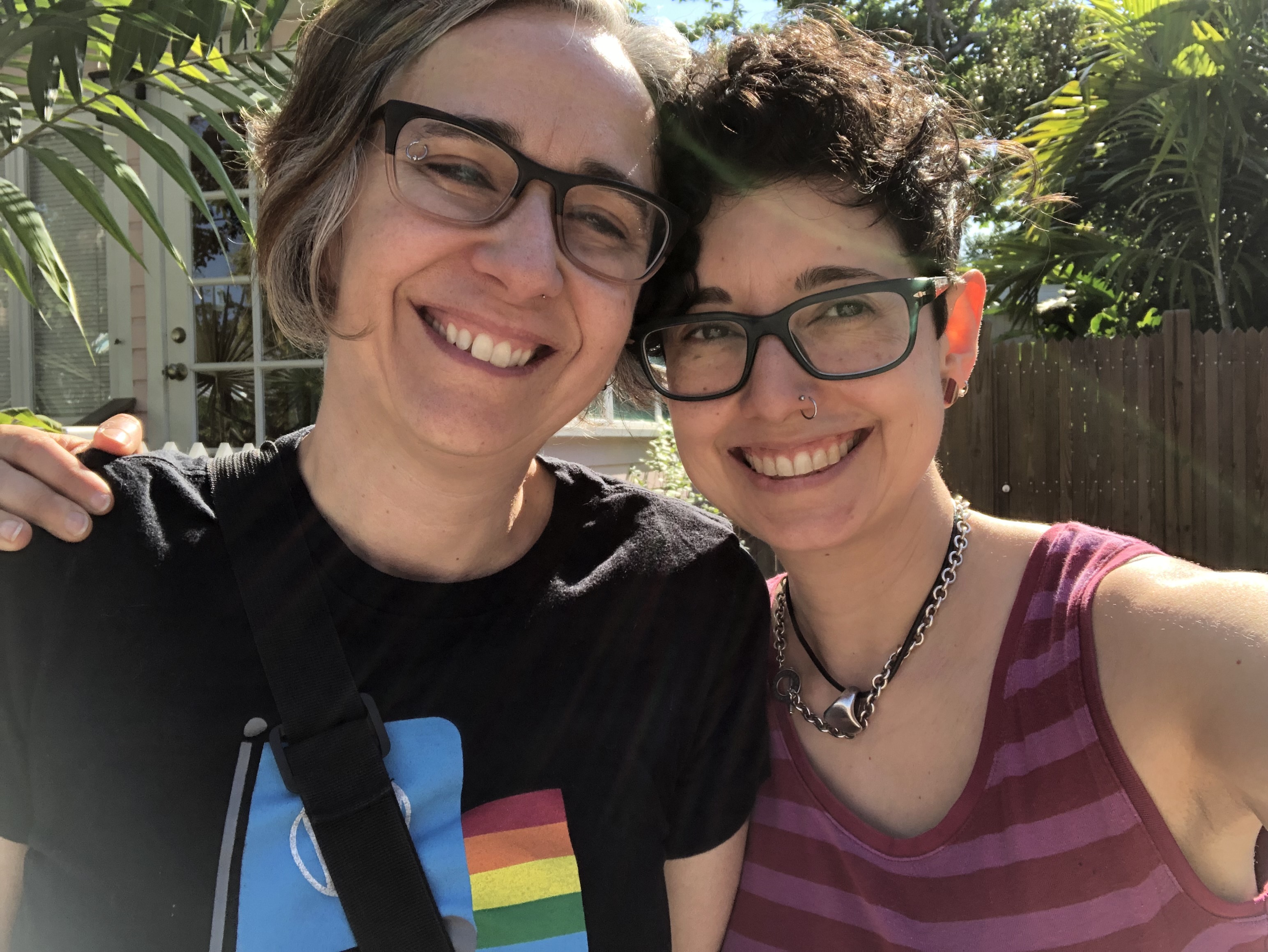 Mia and Tracy stand in a sunny, palm-dense outdoor area in Key West. This photo looks fairly recent. Mia has her short, brown streaked with gray, bob, and Tracy's hair is short and culry on top. They both wear glasses. Tracy jas a mpse ring and Mia an eyebrow ring