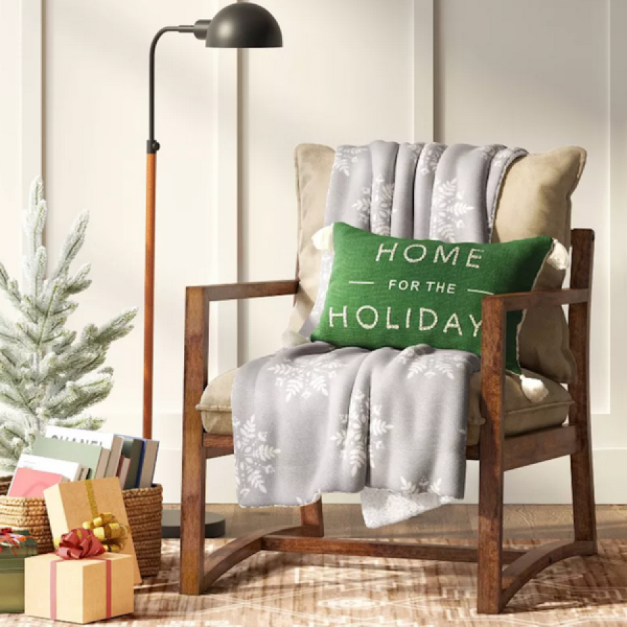 a light gray blanket with white snowflakes draped over a beige and wood chair with a green throw pillow over it. there is a tall brown and black lamp, presents on the floor, and a small pine tree in the corner