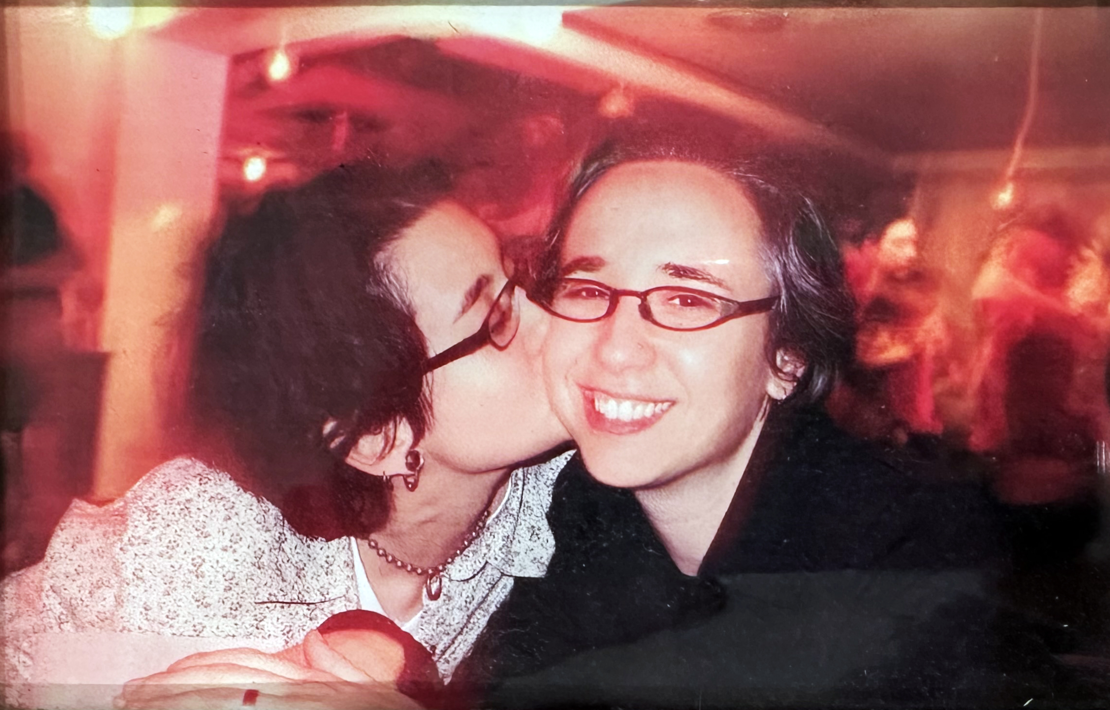 Sometime in the late 90s or early 2000s, judging by the glasses, Tracy kisses Mia's cheek while Mia smiles at an event with blurry red lighting and dancing or moving figures in the background.