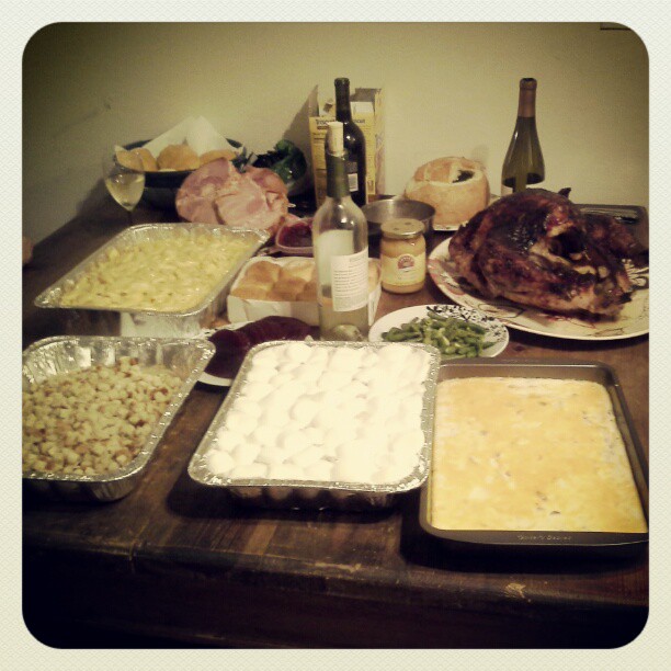 a dark wood table. on it are 3 wine bottles, 1 half full wine glass, a bowl of rolls, a pan of mac n cheese, a pan of stuffing, a pan of sweet potatoes covered in marshmallows, and a pan of cheesy potatoes. There is also a brown glazed turkey