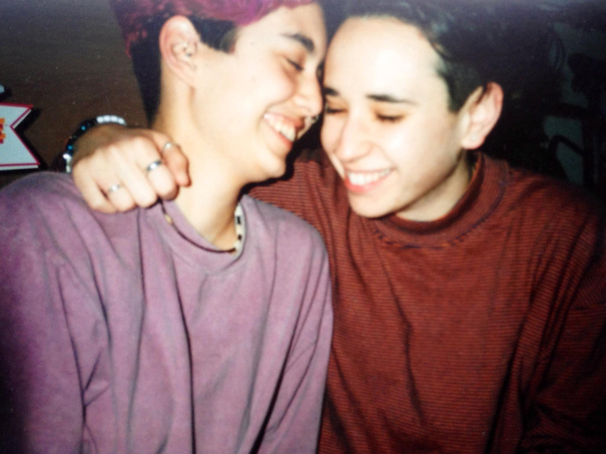 Tracy and Mia embrace in the 90s. Mia's hair is very short and dark. Tracy's is partially dyed pink. Mia is wearing a red long sleeve and a lot of rings and Tracy is wearing a pink sweatshirt and beaded necklace