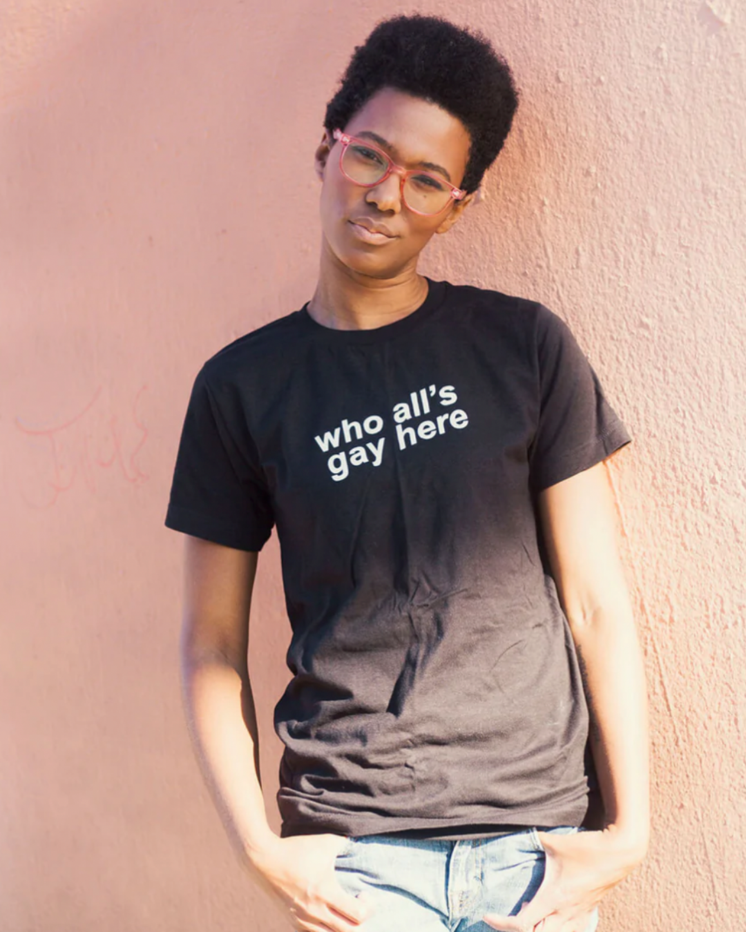 Model Sai is wearing the Who All’s Gay Here Black Tee in size S. She is 5'9" and her bra size is 34B. The tee is black with white sans-serif text that says "Who All's Gay Here." The text is askew at a diagonal in two lines. The design is by Erin Sullivan.