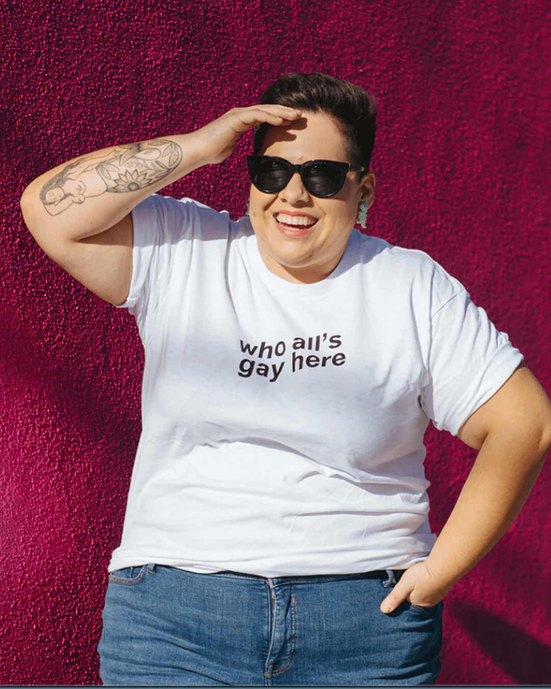 Model Dafne is wearing the Who All’s Gay Here White Tee in size XL. She is 5'1" and her bra size is 44D. The tee is white with black sans-serif text that says "Who All's Gay Here." The text is askew at a diagonal in two lines. The design is by Erin Sullivan.